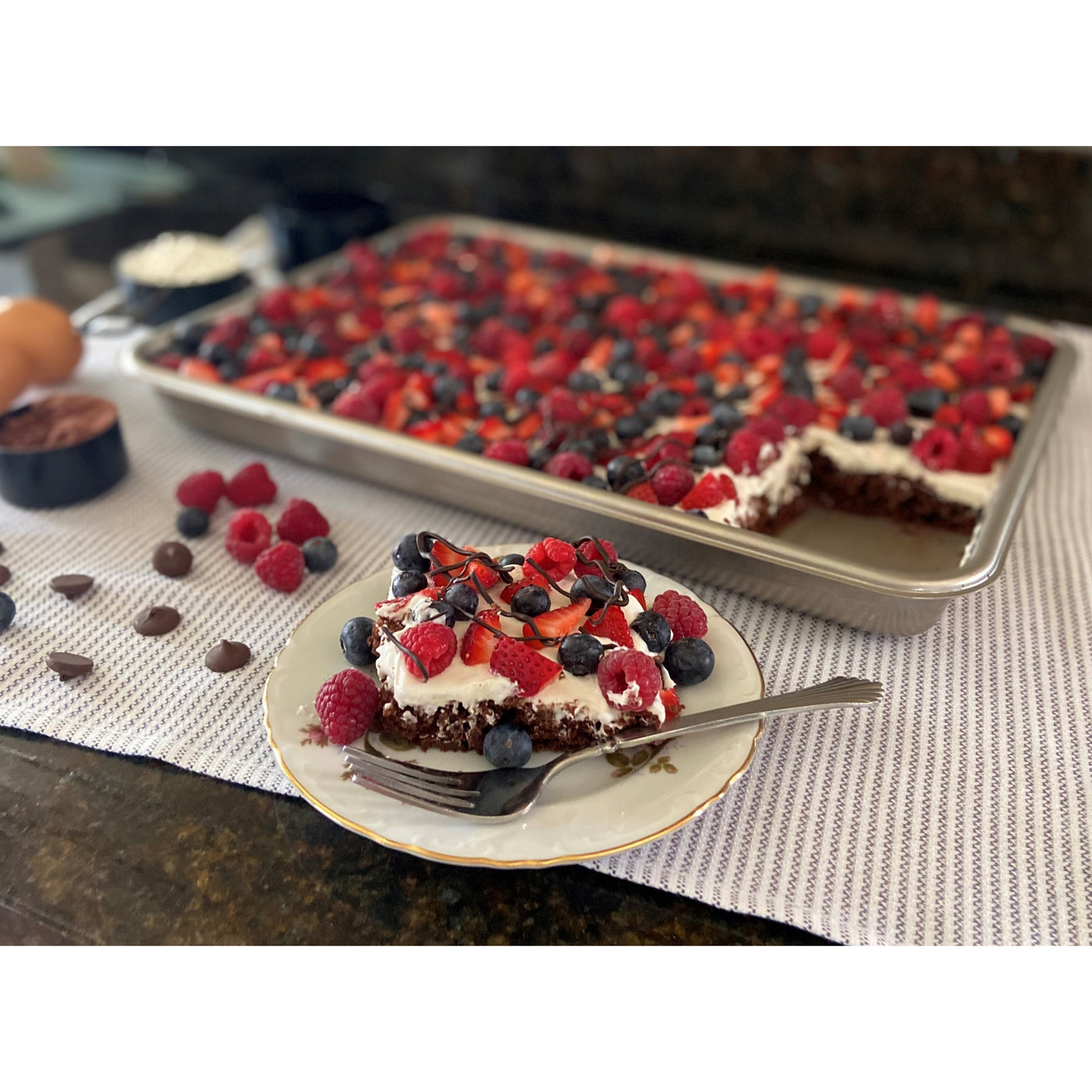 Slice of berry-topped cake on a plate with a larger tray of cake in the background.